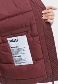 Bordeauxkleurige pufferjack binnenzijde met een gewatteerde textuur, een zijzak met rits en een onderhoudsetiket met zwarte tekst op een witte achtergrond.