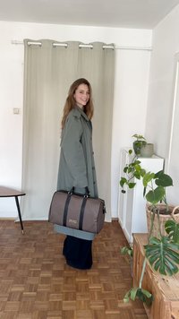 Borsa a duffle marrone a motivo con manici neri, chiusura con zip e un logo dorato, tenuta da una persona in un cappotto verde su uno sfondo neutro.
