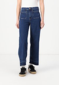Jeans de tiro alto y pierna ancha en denim azul oscuro con bolsillos frontales, costuras visibles y dobladillo sin acabar. Combinados con zapatillas negras.
