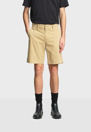 Man wearing beige knee-length shorts, black crew socks, black ankle boots, and a black t-shirt, standing against a plain background.