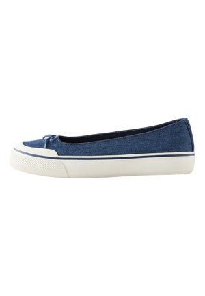 Pieds portant des chaussures bleu slip-on avec semelles en caoutchouc blanches et petits nœuds, associés à un pantalon blanc sur un fond de sol neutre.