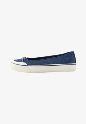 Pieds portant des chaussures bleu slip-on avec semelles en caoutchouc blanches et petits nœuds, associés à un pantalon blanc sur un fond de sol neutre.