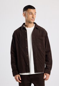 Brown corduroy shirt jacket with a button front, single chest pocket, and a relaxed fit. Textured fabric and collar detail visible.