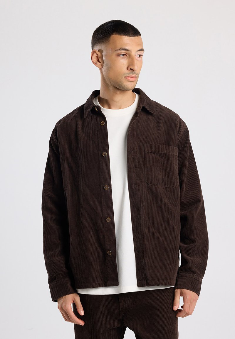 Brown corduroy shirt jacket with a button front, single chest pocket, and a relaxed fit. Textured fabric and collar detail visible.