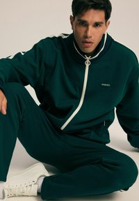 Ciemnozielony dres z wysokim kołnierzem i białym zamkiem błyskawicznym; zawiera białe akcenty na rękawach. Noszony z białymi trampkami. Gładka tekstura.