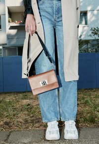 Beżowy trencz, niebieskie dżinsy szerokie na nogawce, białe trampki. Brązowa torba na ramię ze srebrnymi okuciami, czarny pasek zwisa z ręki.