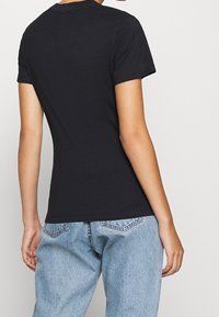 Osoba w jednolitym czarnym T-shircie z krótkim rękawem oraz jasnoniebieskich jeansach z wysokim stanem, stojąca tyłem do aparatu.