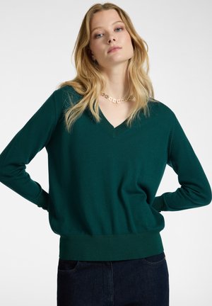 Grüner V-Ausschnitt-Pullover mit langen Ärmeln, gerippten Bündchen und Saum, aus weichem Material, kombiniert mit dunklen Jeans und einer blumigen Halskette.