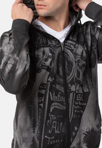 Sweat-shirt à capuche noir et gris, arborant un graphique de moto vintage et du texte. Le matériau est doux, avec une fermeture éclair et des poches.