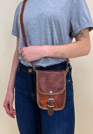 Bruine leren schoudertas met een klep, zilveren hardware en een verstelbare schouderband. Draag je over een grijs shirt en donkere jeans.