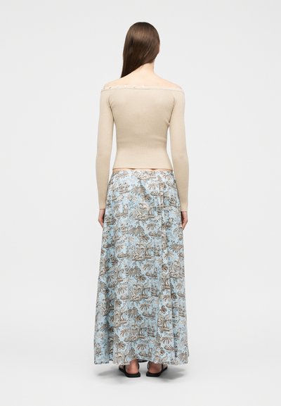 Γυναίκα με μακριά καστανά μαλλιά, φορώντας μπεζ μπλούζα με μακριά μανίκια και άνοιγμα στους ώμους και μπλε μακρύ φούστα με μοτίβα, με την πλάτη στραμμένη, σε λευκό φόντο.