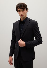 Giacca da abito nera con vestibilità slim, chiusura a un bottone, rever a notched e taschino sul petto; abbinata a una camicia scura con bottoni.