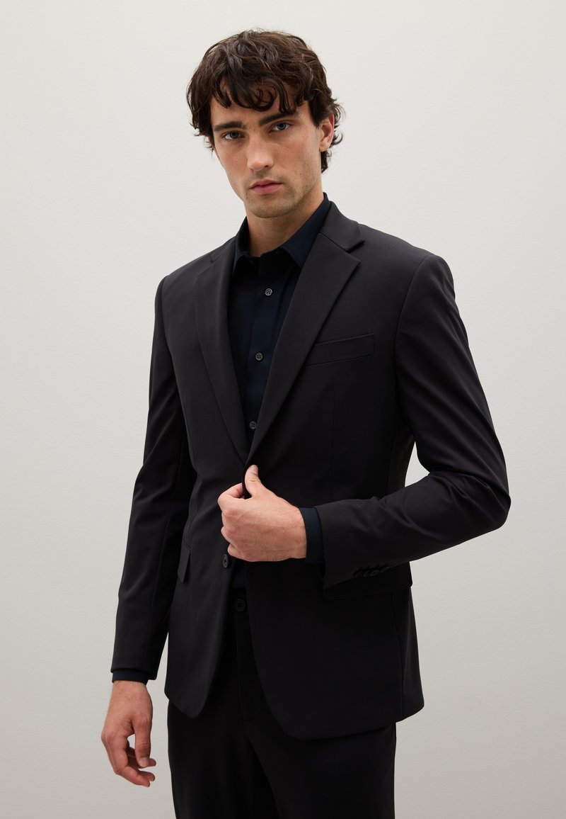 Giacca da abito nera con vestibilità slim, chiusura a un bottone, rever a notched e taschino sul petto; abbinata a una camicia scura con bottoni.