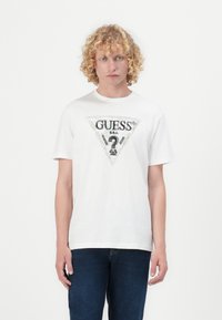 Valkoinen puuvillapaita, jossa on triangelimainen, hopealla koristeltu "GUESS"-logo keskellä, yhdistettynä sinisiin farkkuihin.