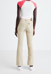 Person som bär beige utsvängda manchesterbyxor och vita sneakers, stående med ryggen mot oss och en kortärmad skjorta i vitt och korall.