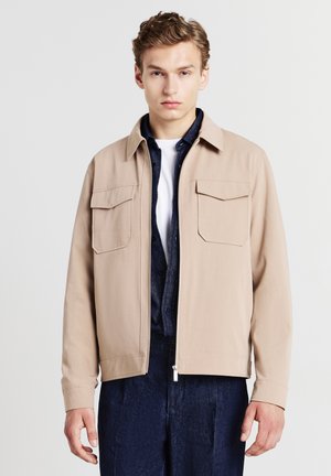 Giovane man in giacca beige con zip sopra una camicia blu scuro e una maglietta bianca, in piedi su uno sfondo chiaro e uniforme.