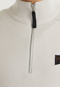 Hellgrauer Reißverschluss-Sweatshirt mit geripptem Kragen, ausgestattet mit einem metallischen Reißverschluss und einem kleinen dunklen Patch mit einem orangefarbenen Logo auf der Brust.