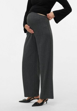 Kobieta w ciąży, ubrana w szerokie szare spodnie i czarne buty na obcasie typu slingback, delikatnie trzymająca brzuch jedną ręką.
