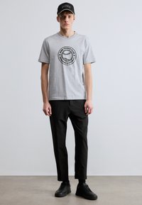 Pilko medvilninio marškinėlių su apvaliu juodu grafinio logotipo tekstu „BOSS TENNIS CLUB“ priekyje derinys su juodomis aptemptomis kelnėmis ir juodais sportbačiais.