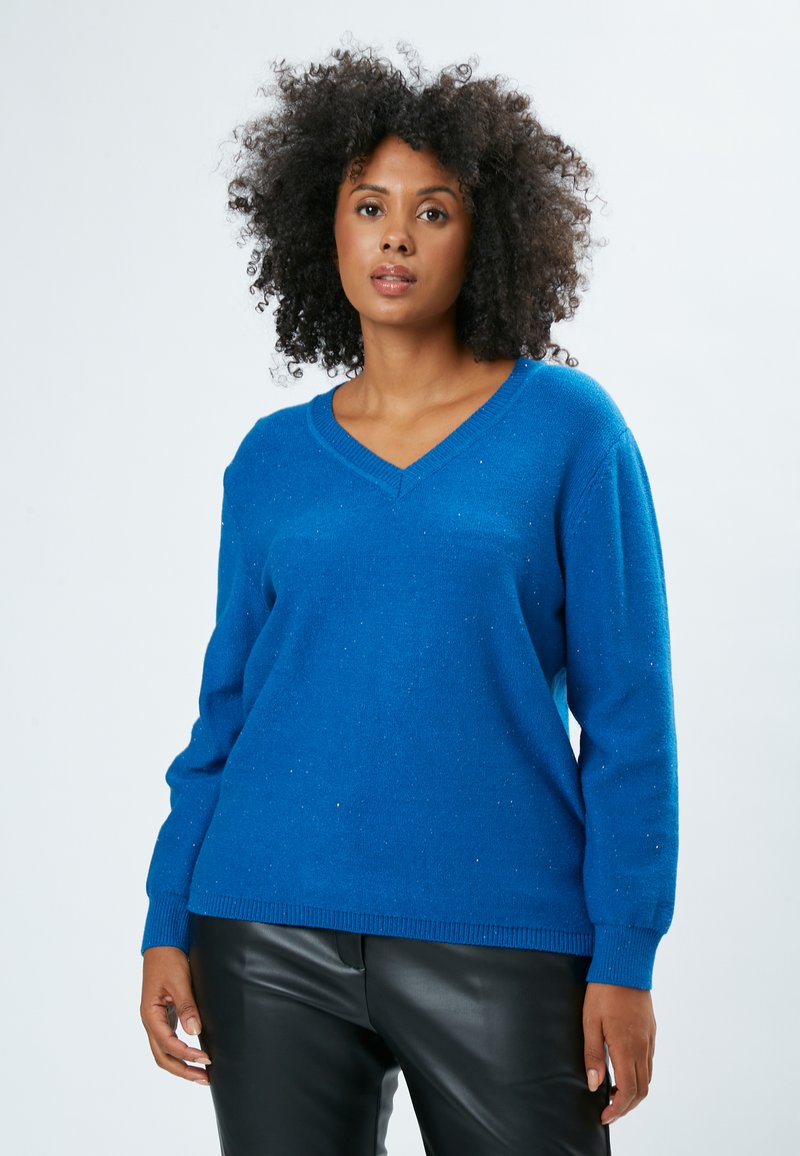 Blauer Pullover mit V-Ausschnitt und funkelnder Textur, langen Ärmeln und gerippten Bündchen. Kombiniert mit schwarzen Lederhosen, vor einem schlichten Hintergrund.