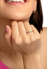 Anillo de piedras preciosas coloridas con una banda delgada, que presenta diversas piedras brillantes en rojo, verde, azul y amarillo. La mano descansa cerca de la barbilla.