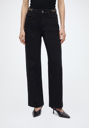 Personne portant un pantalon noir taille haute, coupe droite, avec des détails de ceinture chaîne dorée et des chaussures à talons noires à bout pointu, debout sur fond blanc.