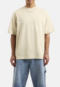 Beżowa, oversize koszulka z bawełny, z krótkimi rękawami, okrągłym dekoltem oraz nadrukowanym logo z przodu. Noszona z niebieskimi szortami dżinsowymi.