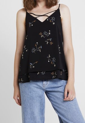 Femme portant un haut sans manches noir à motifs floraux avec fines bretelles et ourlet superposé, associée à un jean bleu clair sur un fond uni.