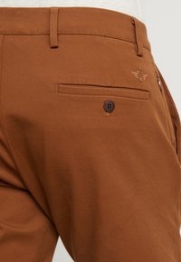 Bruine katoen broek met een achterzak met knoopsluiting en subtiele geborduurde logo details. Zachte textuur en getailleerde pasvorm.