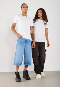 To personer har hvide t-shirts på. Den ene har blå denim shorts med bred ben og sorte støvler; den anden har mørke cargo bukser og hvide sneakers.