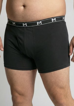 Mannelijk torso en bovenbenen in zwarte boxershorts met een zwarte tailleband voorzien van witte "M"-logo's, tegen een effen lichte achtergrond.