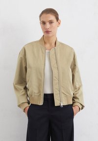 Veste bombardier en beige clair avec une finition lisse, des poignets côtelés et une fermeture éclair à l'avant. Comprend des poches latérales et une doublure matelassée.
