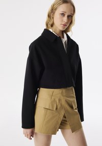 Schwarze Zip-Jacke mit hohem Kragen und weiten Ärmeln, kombiniert mit hellbraunen, hochgeschnittenen Shorts mit Klapptaschen und rohem Saum.