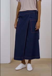 Denim zavojna suknja s velikim prednjim džepovima, kontrastnim šavovima i dugim siluetom. Uparena s jednostavnom majicom i bijelim tenisicama.