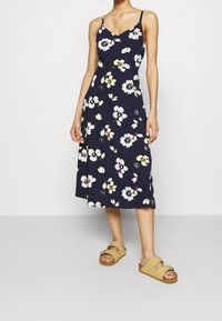 Marineblaues Blumenkleid mit Spaghettiträgern, ausgestattet mit weißen und gelben Blumen, knielang, aus leichtem Stoff. Braune Rutschsandalen.