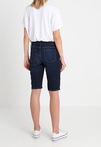 Tmavě modré denimové bermudy s středním pasem, mají zadní kapsy a rovný střih, nošené s volným bílým tričkem a bílými teniskami.