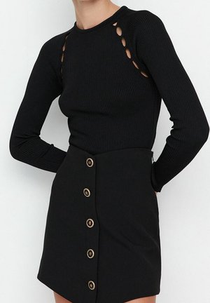 Femme portant un haut noir côtelé à manches longues avec des découpes aux épaules et une jupe noire avec une rangée verticale de cinq boutons bordés d’or.