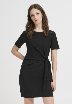 Donna con capelli lunghi e castani che indossa un vestito nero a maniche corte con un nodo laterale, in piedi davanti a uno sfondo chiaro e semplice.