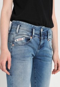 Modré denimové džíny s mírným vyblednutím, s prošívanými kapsami, metalickými akcenty a modelem s logem na přední straně. Nosené s černým topem.