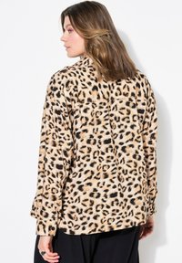 Žena s hnědými vlasy po ramena, oblečená v tričku s leopardím vzorem a černých kalhotách, stojí čelem vlevo s hlavou otočenou na stranu.