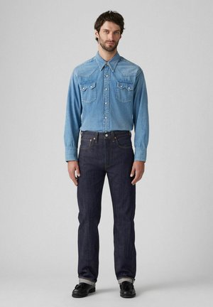 Camisa de mezclilla azul con collar puntiagudo y dos bolsillos en el pecho, combinada con jeans oscuros que tienen costuras en contraste y un corte recto.