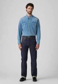Camisa de mezclilla azul claro con dos bolsillos en el pecho, puños abotonados, combinada con jeans de mezclilla azul oscuro. Unos zapatos negros completan el conjunto.