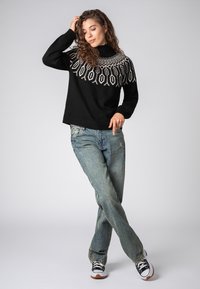 Schwarzer Strickpullover mit weißen geometrischen Mustern, kombiniert mit hellblauen Jeans und schwarzen Sneakers. Schlichtes und lässiges Design.