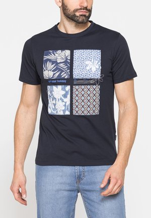 Uomo che indossa una t-shirt blu navy con quattro toppe quadrate con disegni floreali e geometrici, abbinata a jeans azzurri.