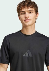 Svart atletisk t-shirt med korta ärmar, rund hals och texturerad logotyp. Slät tyg med en lätt lyster och en modern, åtsittande design.