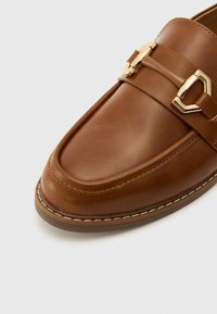 Mocassin en cuir marron avec une texture lisse et un détail de sangle en métal doré. Bout arrondi, accents cousus et semelle en caoutchouc.