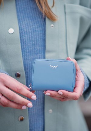 Compact blue leather wallet with a smooth texture, white logo on the front, and a zippered closure, held in hands against a pastel outfit.