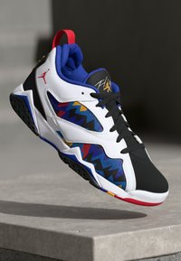 Scarpa da basket bianca, nera e blu con motivi zigzag colorati, linguetta rossa e lacci neri, sospesa sopra una superficie di pietra grigia.