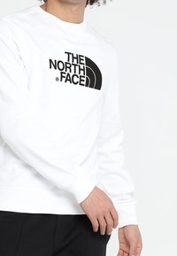 Person som bär en vit sweatshirt med "THE NORTH FACE"-logotypen i svart på bröstet, tillsammans med svarta byxor, mot en helt vit bakgrund.