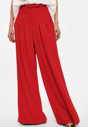 Donna che indossa pantaloni rossi a vita alta e gamba ampia con pieghe frontali e cintura rossa coordinata, abbinati a un top bianco.